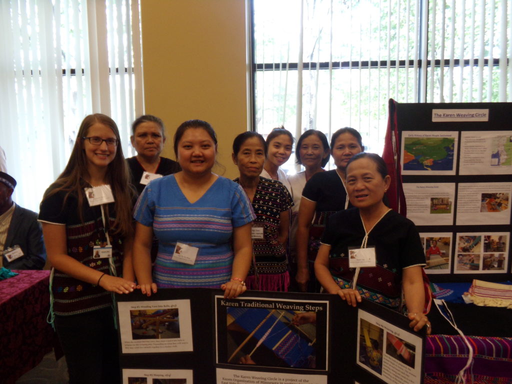 Women in the Karen Weaving Circle at the Food Group event posing in front of weaving table and posters