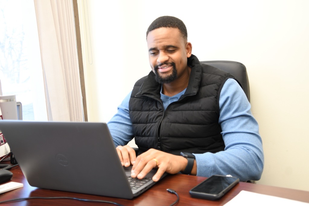 A man sits at a desk and works at a computer while smiling.