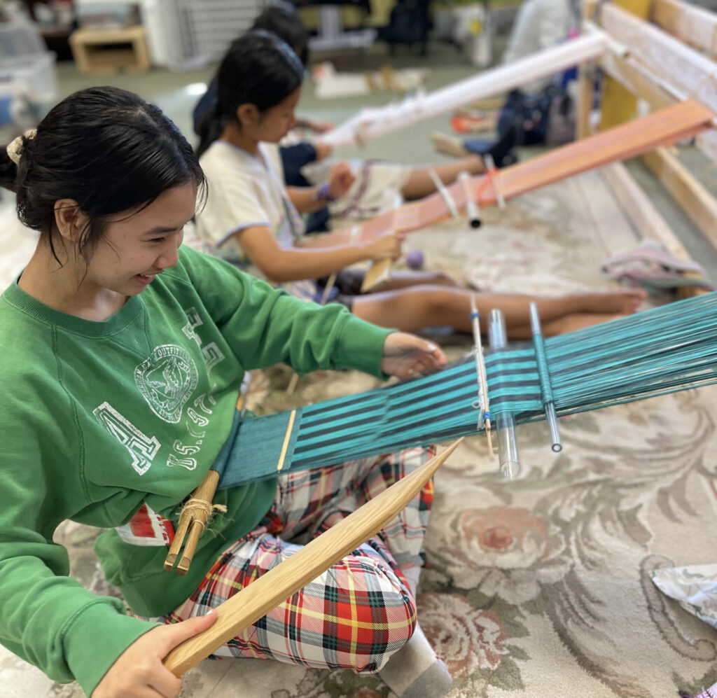 Youth sit on the ground indoors and practice backstrap weaving on a traditional Karen loom.