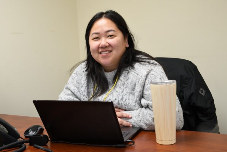 KOM Staff, Lisa Khang, sits at her desk in the office and smiles to the camera.