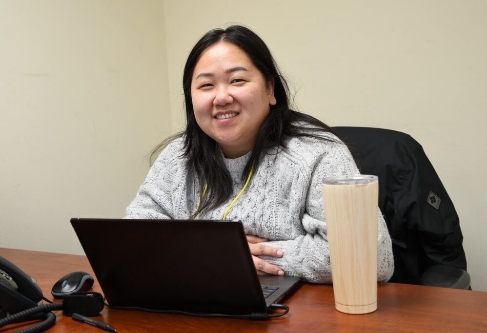 KOM Staff, Lisa Khang, sits at her desk in the office and smiles to the camera.