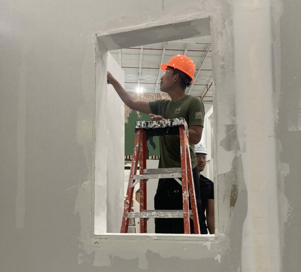 A man paints an unfinished wall while standing on a ladder wearing a construction hard hat.
