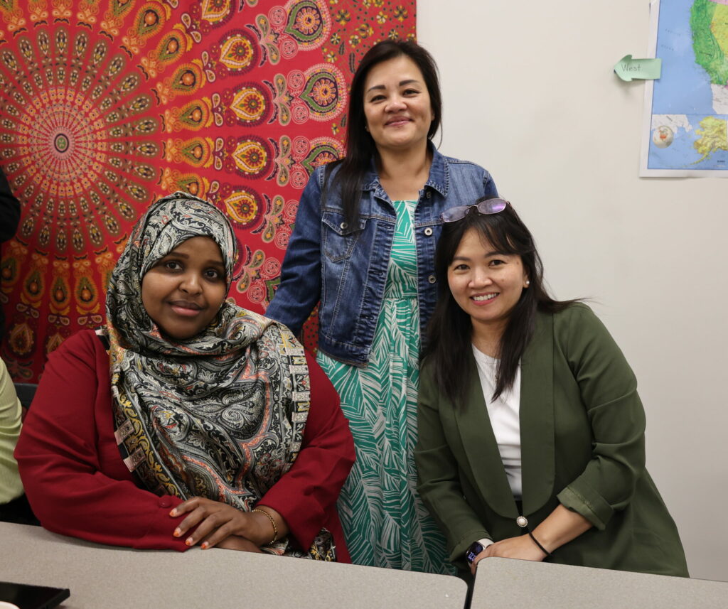 Three adult students pose for a photo, two sitting and one standing, in front of a colorful background wall decoration.