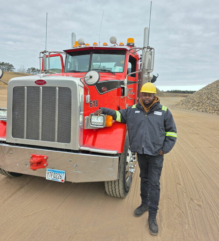 A man dressed in work clothing and a hard hat poses next to a large truck after completing a Commercial Driver's License certification program.