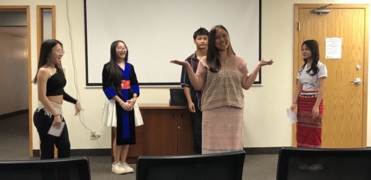 Five youth stand at the front of a room, with one student standing in the center, performing their plays.